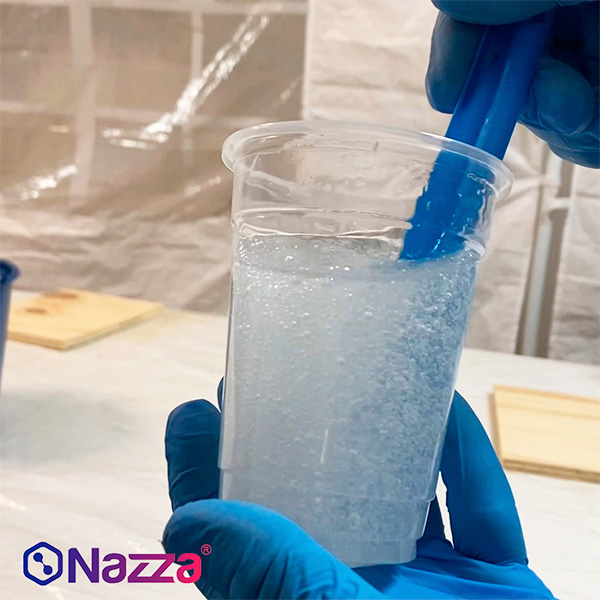 vaso de plástico transparente con resina epoxi recién mezclada, en el que se aprecian numerosas burbujas de aire. Unas manos con guantes azules sujetan el vaso y una espátula de mezcla, removiendo el contenido. La escena transmite el momento inicial del proceso de aplicación, destacando la importancia de una mezcla cuidadosa para evitar la formación de burbujas y conseguir un acabado cristalino en trabajos de coladas profundas o capas gruesas.